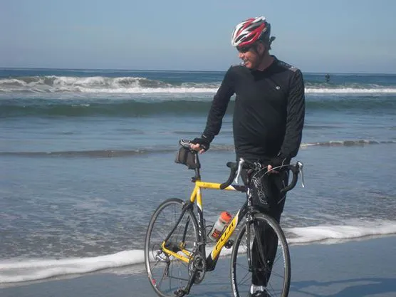 Michael Breu, a white male, in black cycling gear and helmet standing next to a yellow racing bike on the beach with the ocean in the background